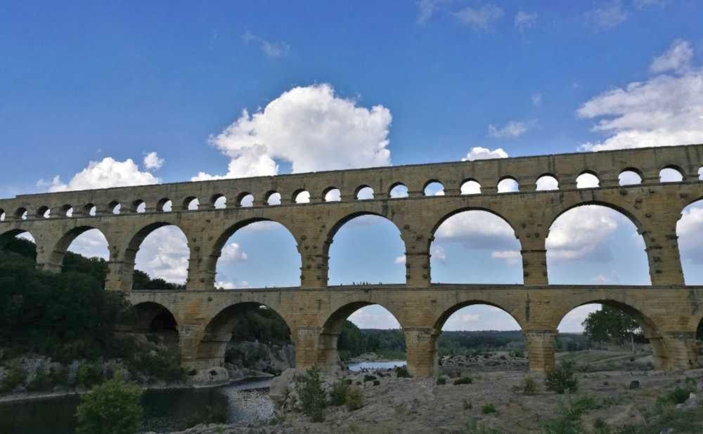 A view of the Pont du Gard from a bit down the riverside. 3 rows of columns on top of each other. The bottom and middle rows are large rounded arches. The top row is made up of much smaller rounded arches, and far more of them too.