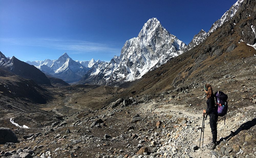A hiker stands on a rocky trail in Sagarmantha National Park. She carries a backpack and leans on two walking sticks as she looks at the scenery: a view of a rocky, snow-capped mountain peak.