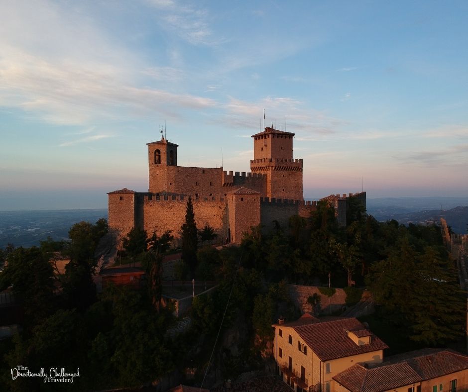 Seen from one side near sunset, the walls of the castle at the top of Mount TItano are crenellated and inside them are two square towers, with walls betweeen them, also crenellated.