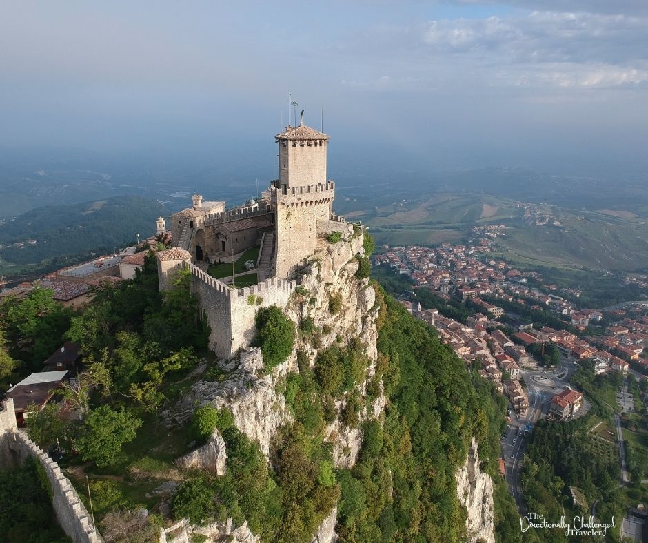 The castle and its walls of stone, perched dramatically on a small peak, with a huge view in the distance. Far below are the roofs of a town. 