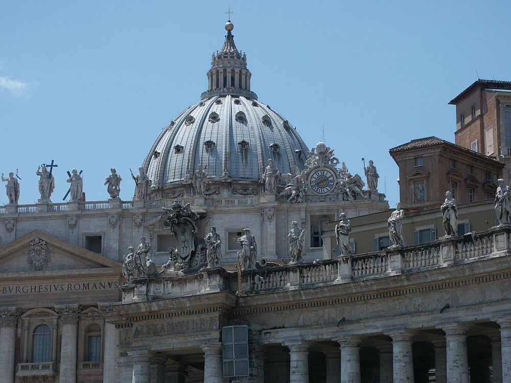 Photo of just one part of the much larger St. Peter's Basilica: the roof is domed, with a point at the top center. Along the edges of the roof at the base of the dome are statues of saints and a very ornate clock. A lower part of the building at an angle to the right is also lined with life-sized saints.