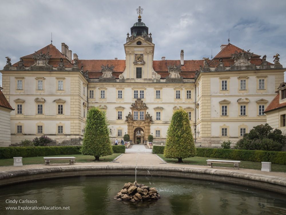 View of Valtice Castle: white stone, about 4 stories tall and symmetrical, with a small tower over an ornate central archway and wings extending forward on both sides of the central archway. In front, a round fountain.