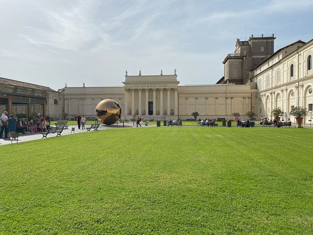 An open, grassy area in Vatican City with a modern, shiny sculpture in it, surrounded by more classical-style buildings.