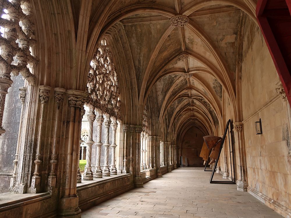 A view along a very long cloister: gothic vaulted ceiling above, plain wall to the right, except where the pillars protrude that support the ceiling, and on the left, the pillars that support the window openings to the cloister's courtyard. These have complicated filigree work on the pillars and on the arches they support.