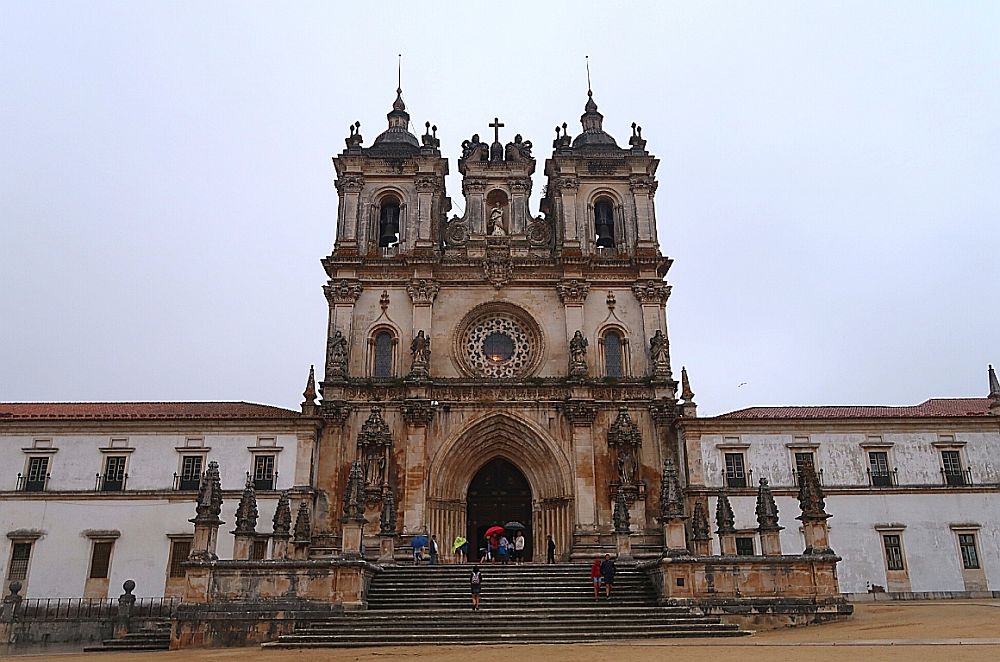 A very elegant central entrance: stairs up to an arched doorway, a rose window above that, rows of statues on the ground floor and above the door, three square towers on top, all with detailed carved ornamentation. The rest of the building is much smaller and extends on both sides out of the picture, two stories tall with simple rows of windows.