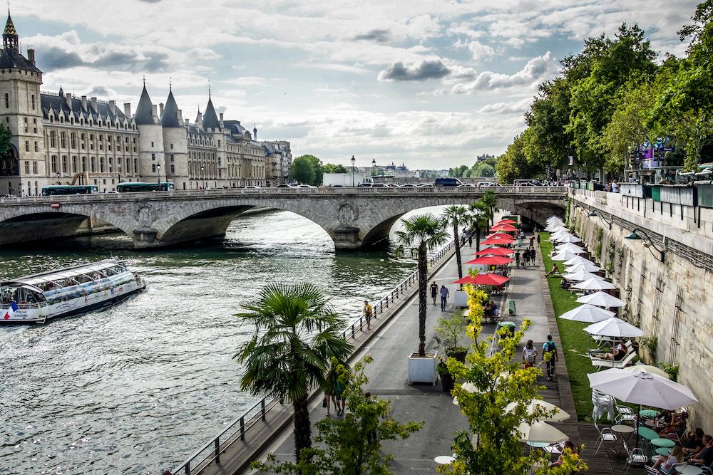 A view along the Seine seen from one bank. Below, lower than street level, is a bank with 2 rows of cafe tables capped with umbrellas. Between the cafe tables and the water is a path with people walking and biking, and a railing along the water. A tourist boat passes under a bridge over the river.
