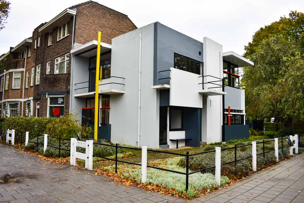 The Rietveld Schröderhuis is all flat surfaces in squares and rectangles. Two stories tall, made of concrete, painted in large areas of gray and white, with a few small vertical and horizontal elements in red and yellow. This view is from the corner, so two sides are visible, both with small balconies.