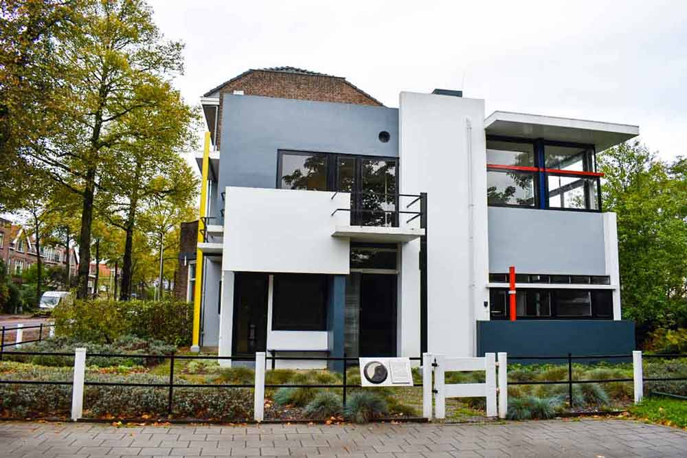 The Rietveld Schröderhuis is all flat surfaces in squares and rectangles. Two stories tall, made of concrete, painted in large areas of gray and white, with a few small vertical and horizontal elements in red and yellow. This view is from The front: a large white vertical panel in the center, a white horizontal panel on the left and grey on the right, large windows.