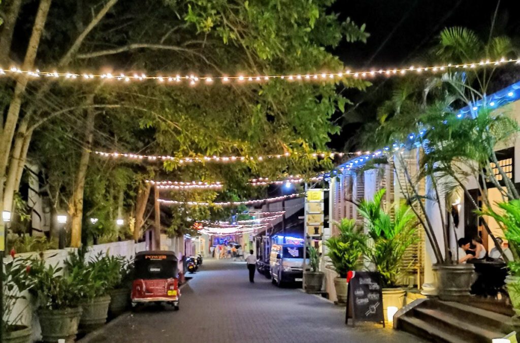 A view of a street at night in Galle Fort: fairy lights stretch across the street, green trees extend over the street, shops on the right and a wall or fence on the left. 