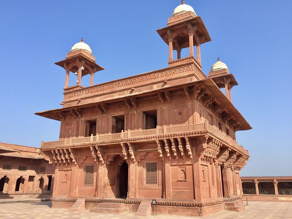 A square building, 2 stories tall, of reddish stone, with a small cupola tower on each corner.