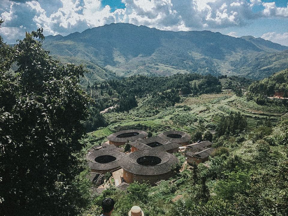 View of some Tulou from above, with mountains in the background. The tulou are 4 round shapes with one that is square.