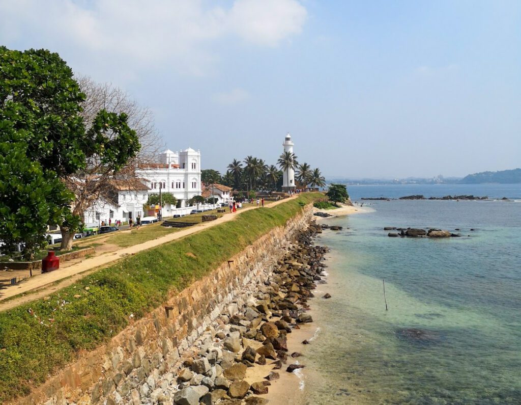 A view down the coast at Galle Fort: edged by a wide wall with a path along its top, some white colonial style buildings along the wall, and a lighthouse in the distance.