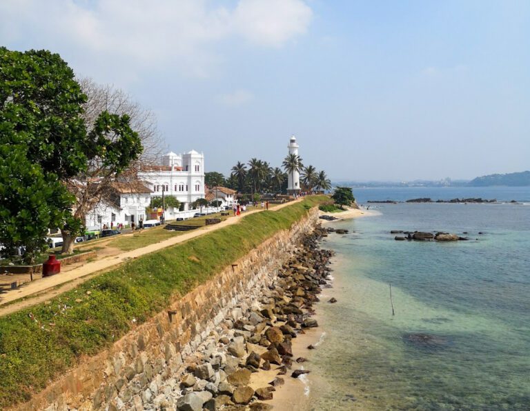 A view along the water with some white colonial buildings in the distance and a lighthouse beyond them.