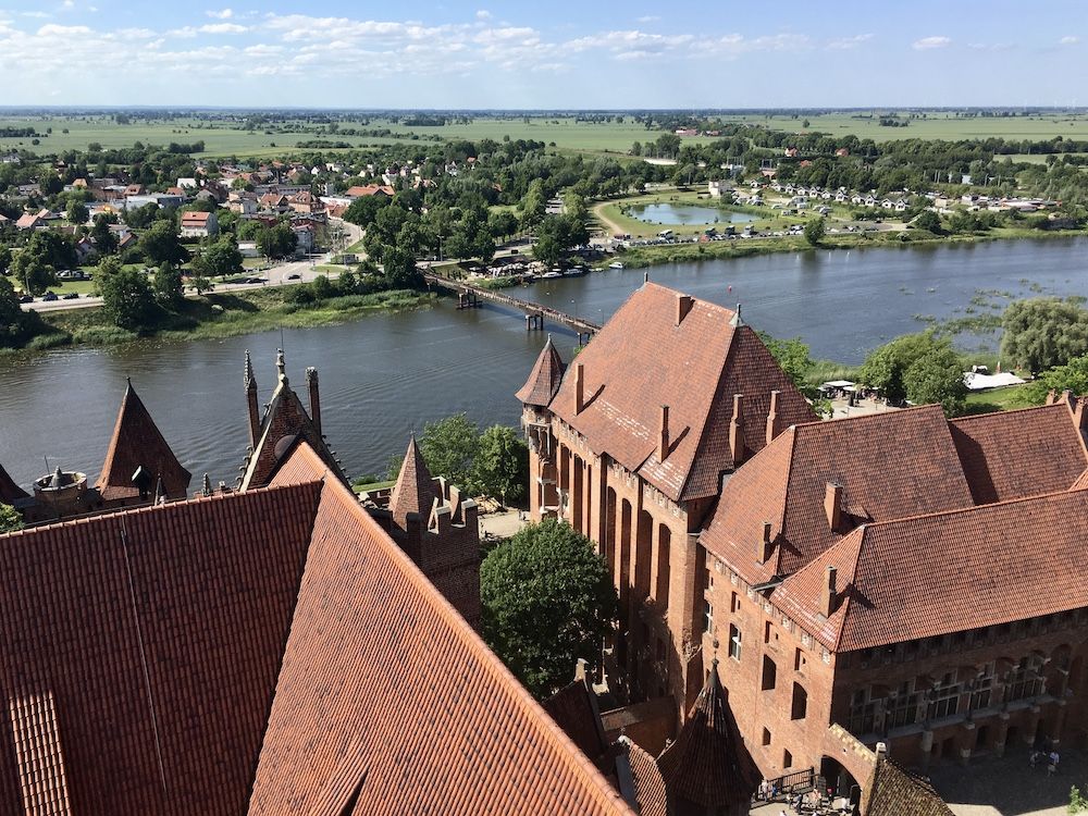 View from a high turret of Malbork Castle looks down on parts of the castle itself and over a river to the countryside.