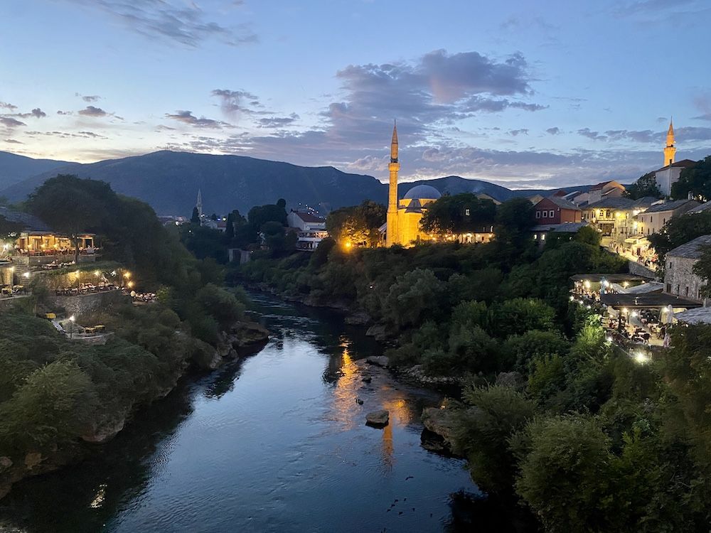 Nighttime view down the river, lights of the town of Mostar on both sides, a mosque on the riverbank whose minaret is lit up.