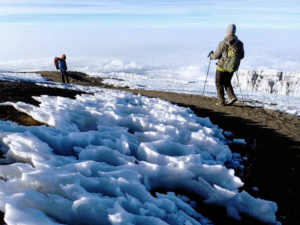 Near the summit of Mount Kilimanjaro, two people stand, bundled warmly and wearing backpacks, and look at the clouds below them. Snow covers much of the ground around them.