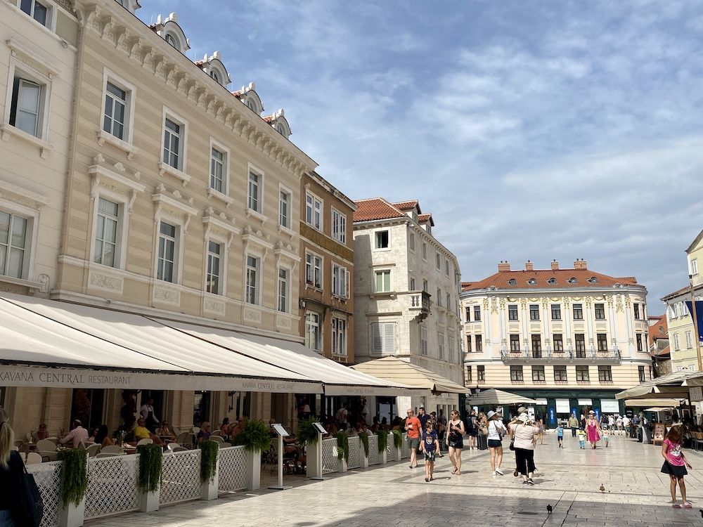 A plaza in Split with elegant buildings along the sides and open air cafes on the ground floors under awnings.