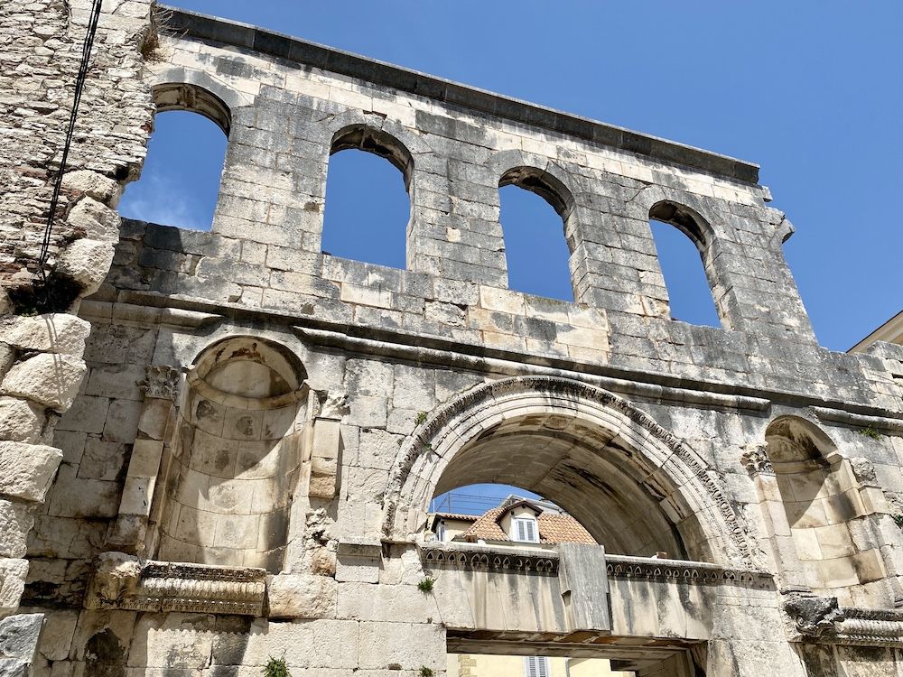 A piece of Diocletian's Palace ruins: A large archway below with empty statue niches on either side, a row of windows above, also arched, with just sky behind them.
