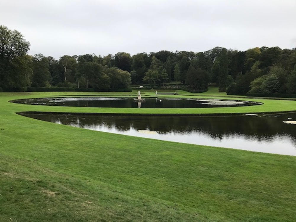 Green lawns around a round pond with a fountain in its center at Studley Royal Park.