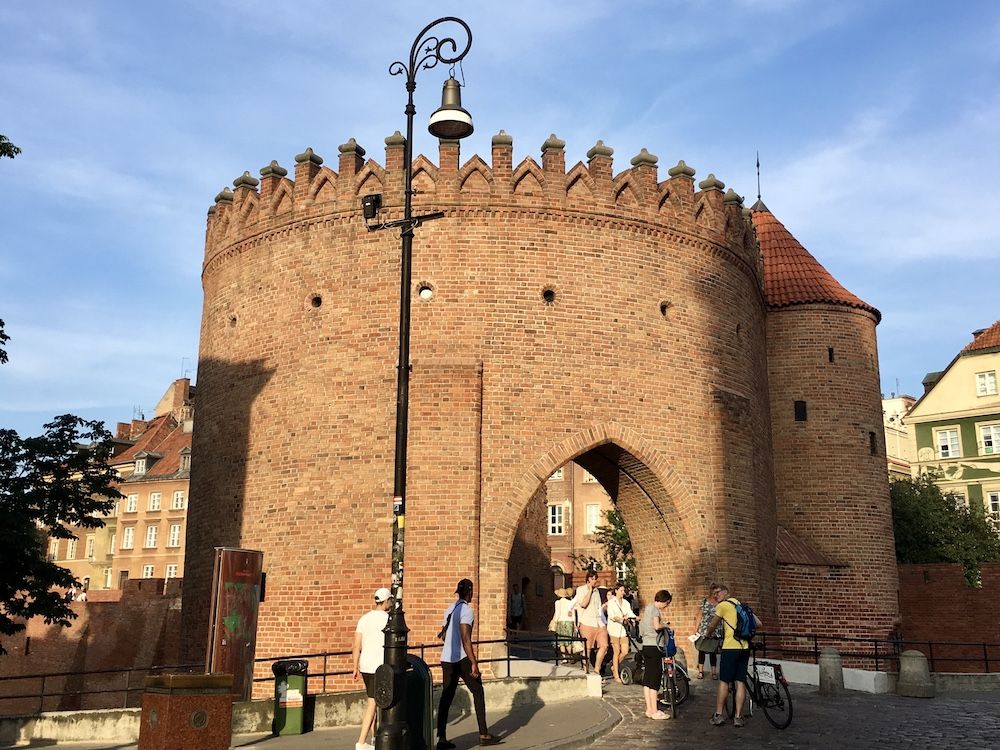 A medieval city gate: brick and round, with an arch through its center and a round tower on its far side. Crenellations around the top.