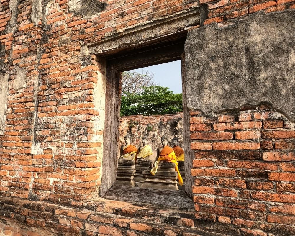 A wall of a building made of red brick, with a window in the middle. Through the window a line of statues are visible, each a bust of a person. The statues have been draped in yellow and orange "clothes."
