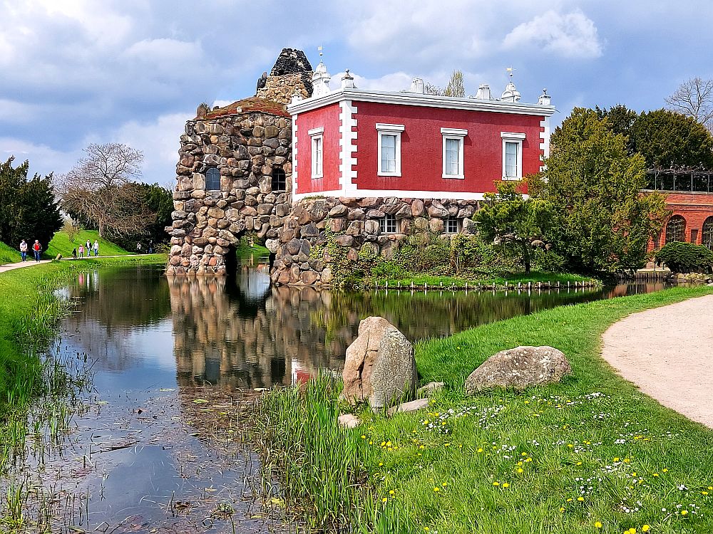 A structure made of large stones, half on land, and half built with arches over a small pond. While most of it is raw stone, one room-sized piece on top is finished and painted bright red with white around the windows.