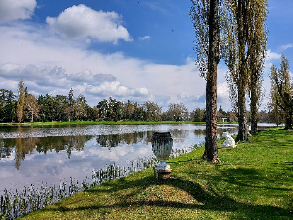 A small lake with trees on near and far side, and a sprinkling of objects: an urn, statues, etc.