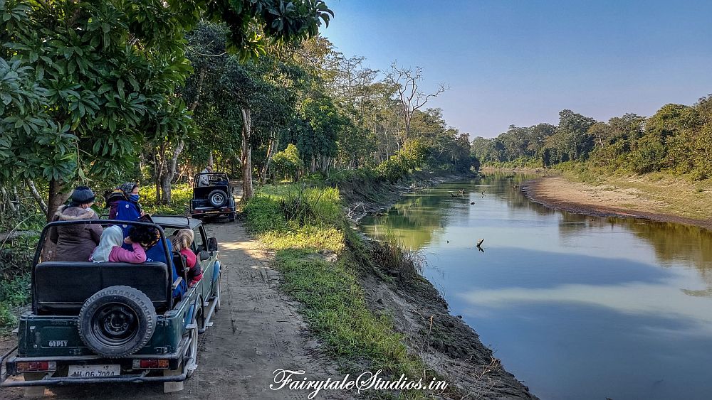 The river on the right, a dirt road along it straight ahead, with two open-top jeeps on the road, each filled with tourists. At Kaziranga National Park.