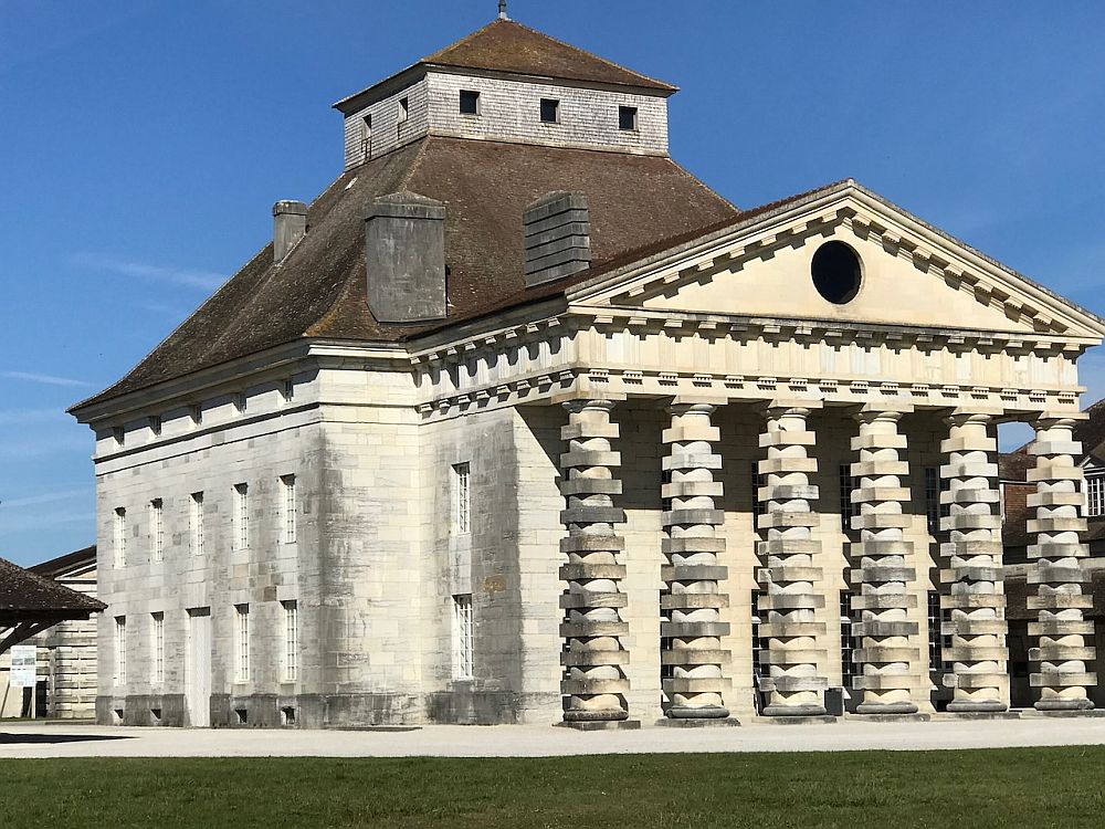 A rather grand white-brick building with a row of 6 oddly-shaped pillars supporting a triangular pediment.