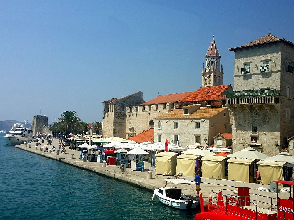 A view of Trogir from the water: market stalls on the quayside, stone buildings behind them, and a piece of a crenellated wall, and a church tower peeking up beyond them.