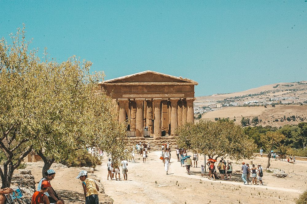 An open area dotted with tourists and a few trees, looking toward the front of a temple with Doric columns holding up a triangular pediment.