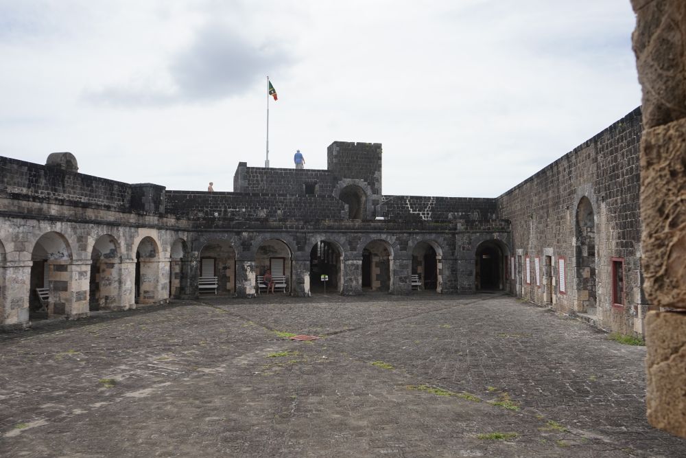 A paved courtyard surrounded by a colonnade of arches in stone, with an upper story for one section, where people are visible.