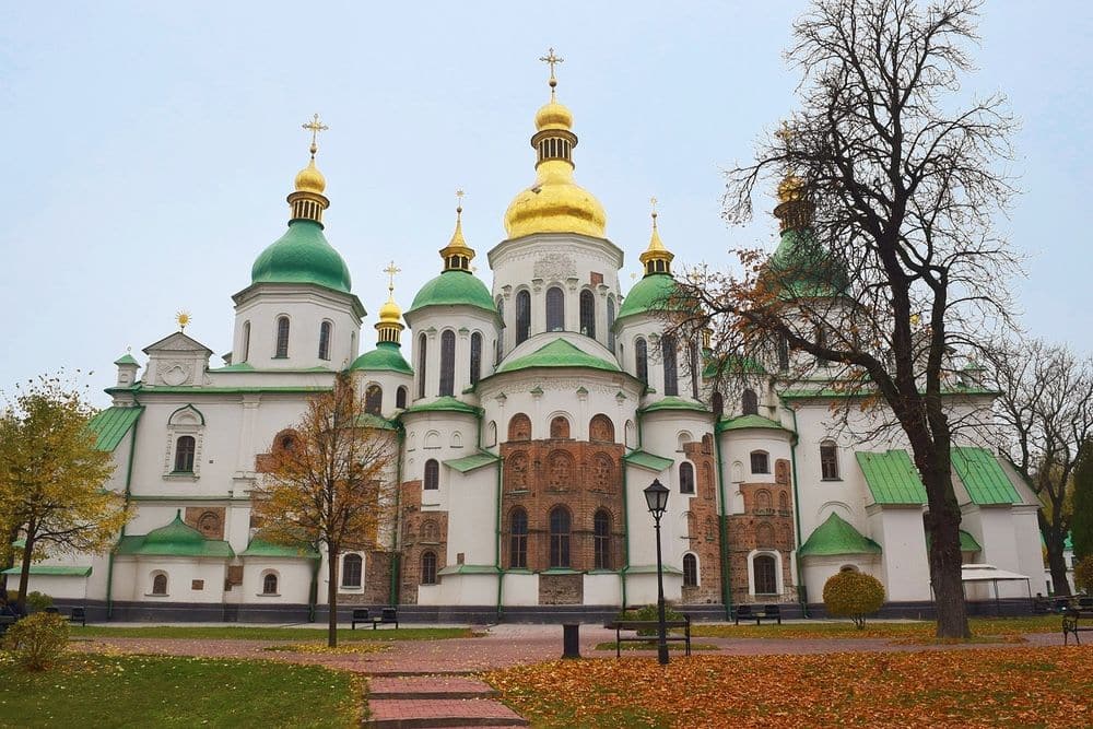 A centered view of Saint Sophia cathedral: sides are mostly white with arched windows. Roofs are green and domed, except the central one which is gold.