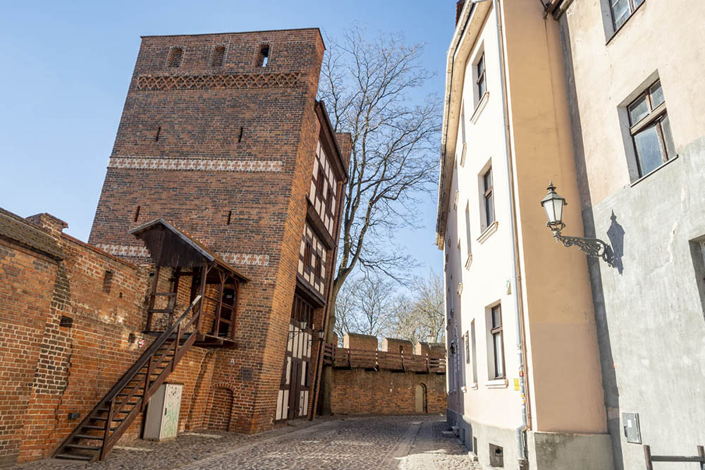 A medieval street: simple houses of 3-4 stories on the right, and a building on the right with brick sides and half-timbered front, which leans visibly over the street.