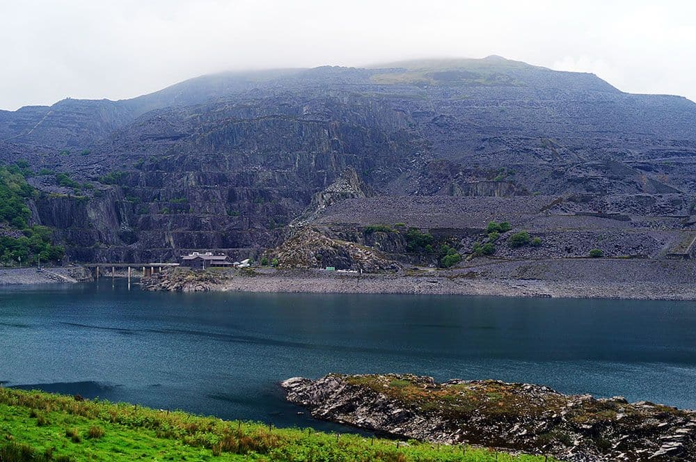 Across a small lake, a mountain looms gray. Its face it cut in large steps until almost the very top of the mountain.