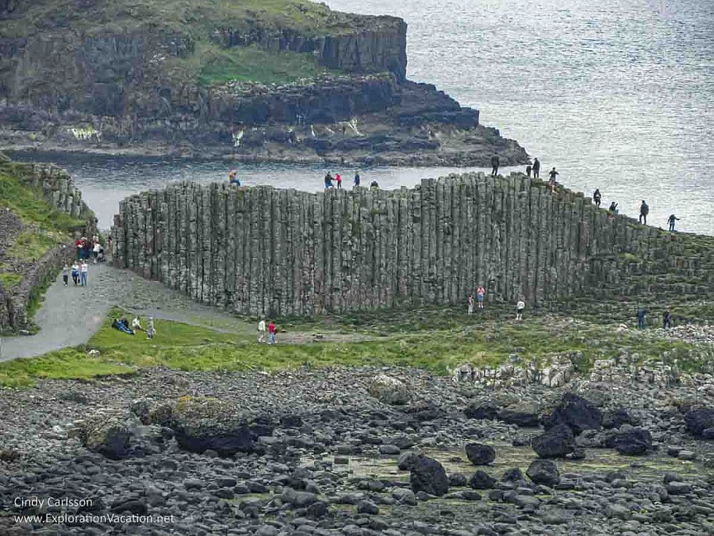 A view of Giant's Causeway from a distance: a wall of stone on the edge of the water with vertical columns, people walking on top of it.