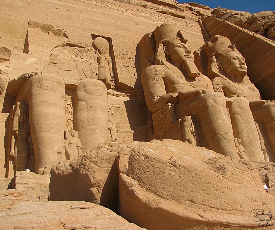 Enormous statues carved out of a rock cliff depict, in this photo, 3 large sitting figures of Egyptian gods. The left-hand one is quite damaged, with only the legs remaining, but the other two are intact.