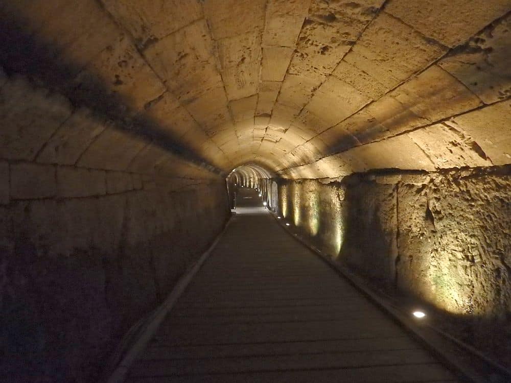 A view down part of the tunnel: straight and with stone walls and arched stone roof, dimly lighted by occasional lights along the floor.