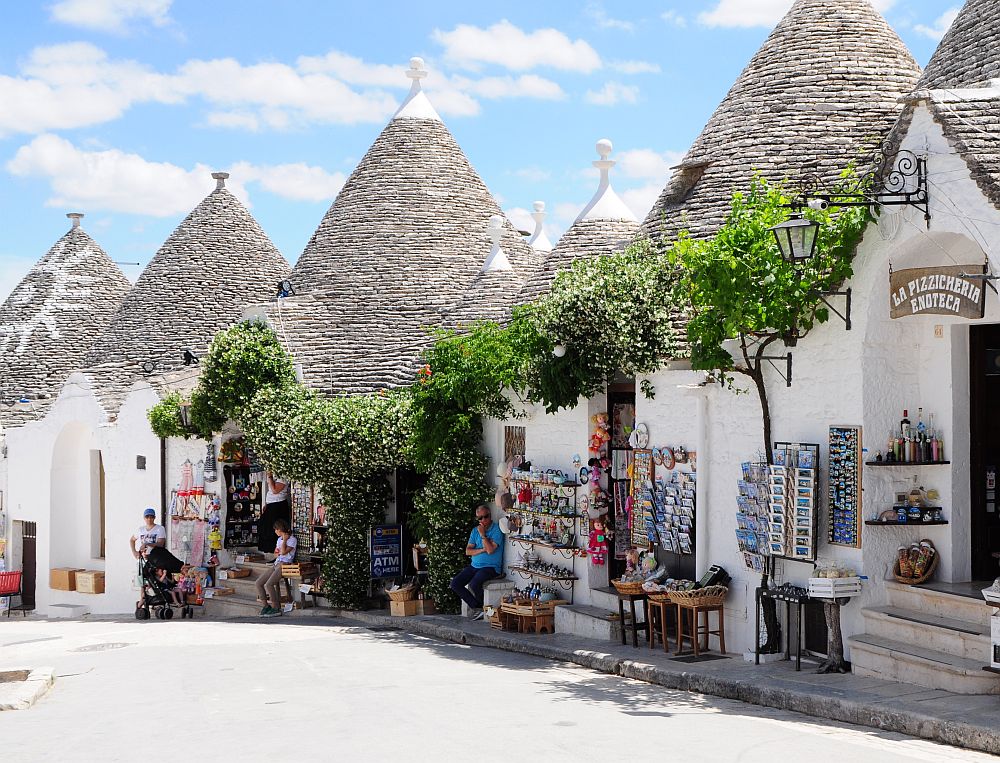 Trulli of Alberobello: A row of white houses with gray slate roofs in conical form. People selling souvenirs in front of them.