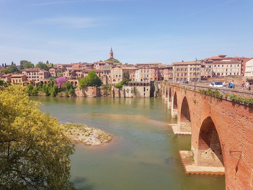 A view from across the river at the Episcopal City of Albi. On the right, a red-brick bridge with arches. On the far bank, many low-rise brick buildings with the steeple of a church visible behind them.