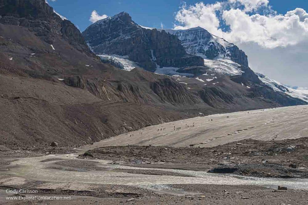 Canadian Rocky Mountain Parks: The glacier is a rounded brown mass, with people walking on it. Behind is a snow-topped mountain.