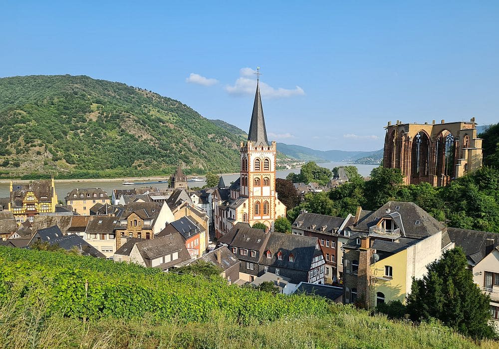 A cluster of buildings with a church in the center, backed by the river and, behind that, hills on the other side of the river.