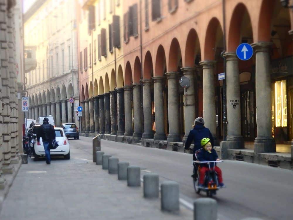 A woman rides a bike down the street, with a child on the back. She passes a row of buildings with porticoes with round pillars and rounded arches.
