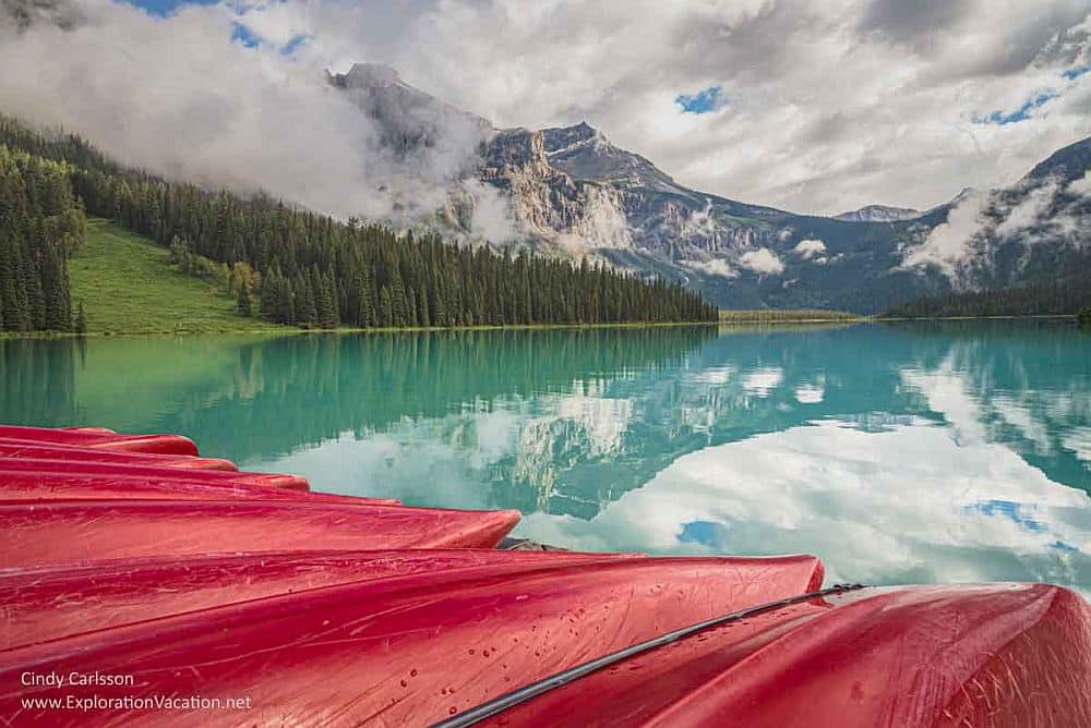 A row of red kayaks waiting on the shore of Emerald Lake - still water, bright greenish-blue, with a background of snow-topped mountains.