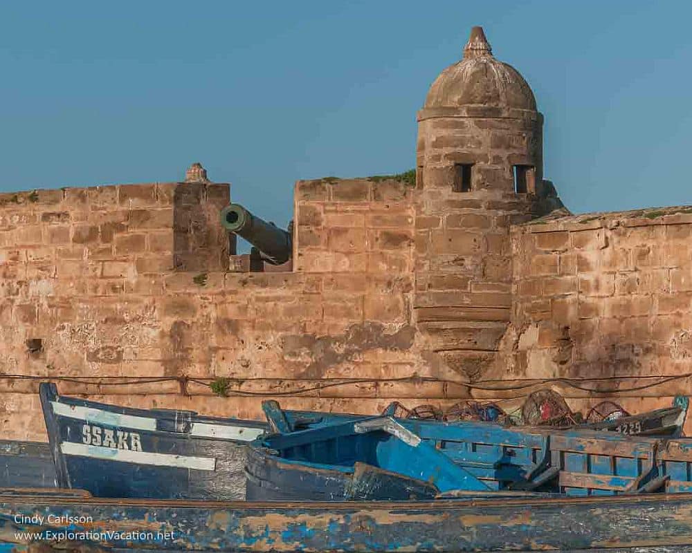 The wall of the Medina of Essaouira's fortification: an opening shows a cannon pointed outward, and a guard post rises next to it. In front of the wall on the water's edge are blue-painted fishing boats.