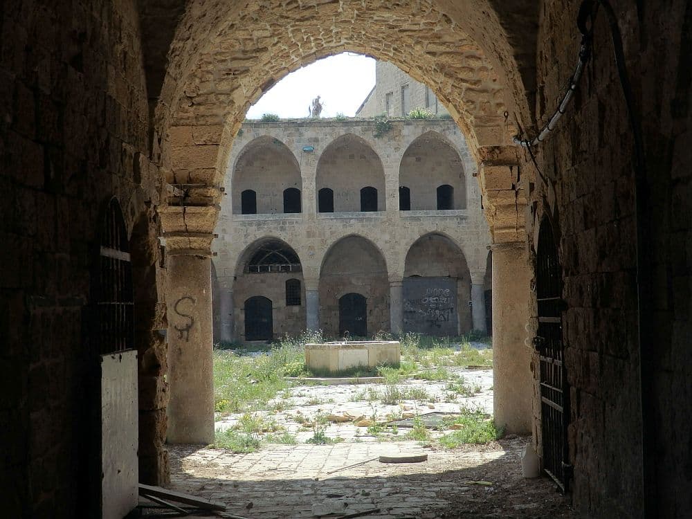 Looking through an archway into an open courtyard, weedy with a disused fountain in the center. On the opposite side, a row of arches supports an upper walkway, which has a roof, also supported by a row of smaller arches.