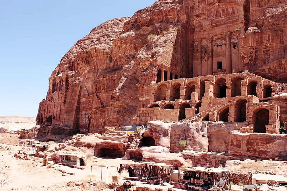 A view of a high cliff with, along its base, a series of tombs with arched entryways, plus some buildings or tombs on the valley floor in front of them.