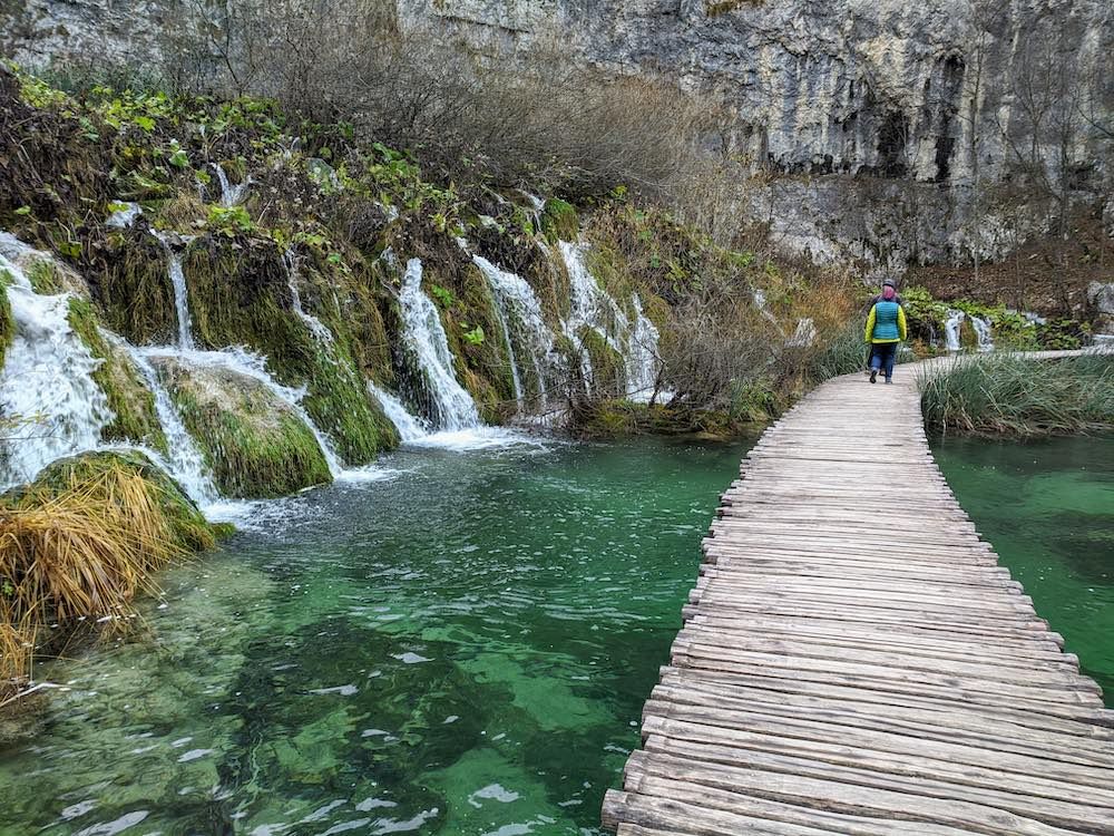 A person follows a boardwalk along the right side of the picture passing, on the left, a series of waterfalls into a pool of clear water.