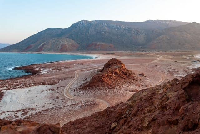 A view from up high over red-sand beach with a small hill in the middle of it, and larger mountains in the background.