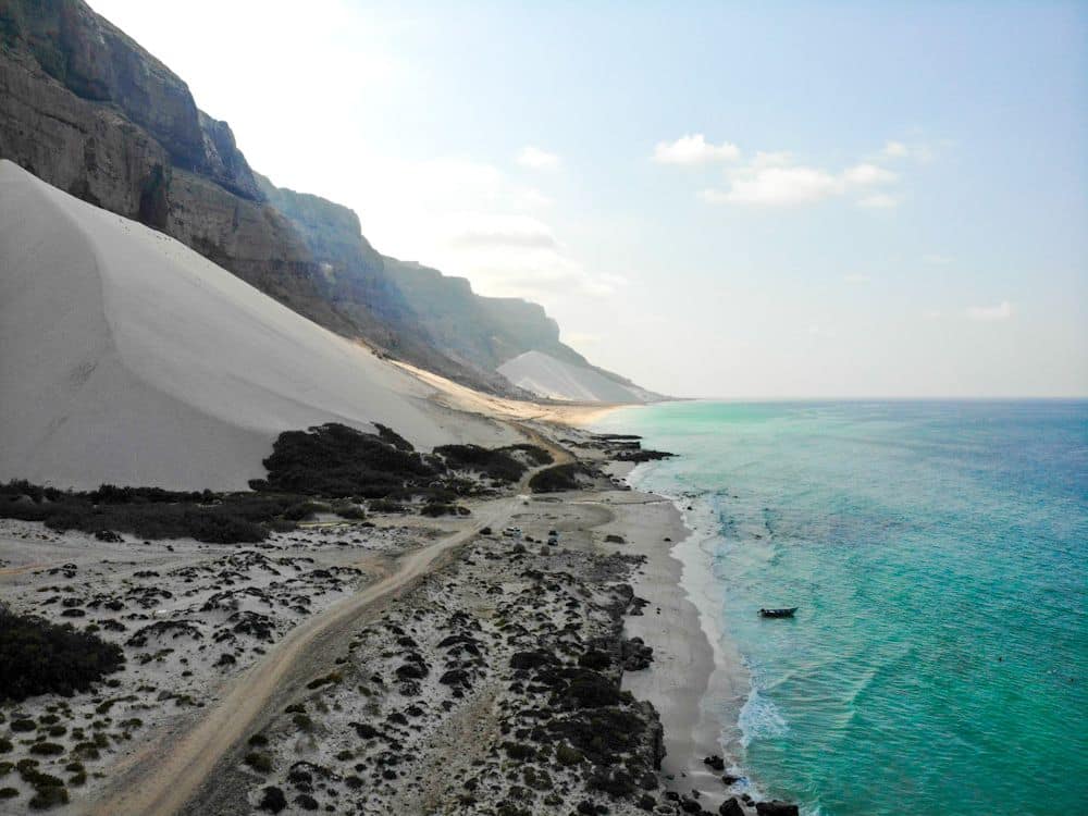 Looking along a shoreline, steep mountains on the left, sand dunes between them and the water's edge. A very small looking boat is visible near the edge of the land.